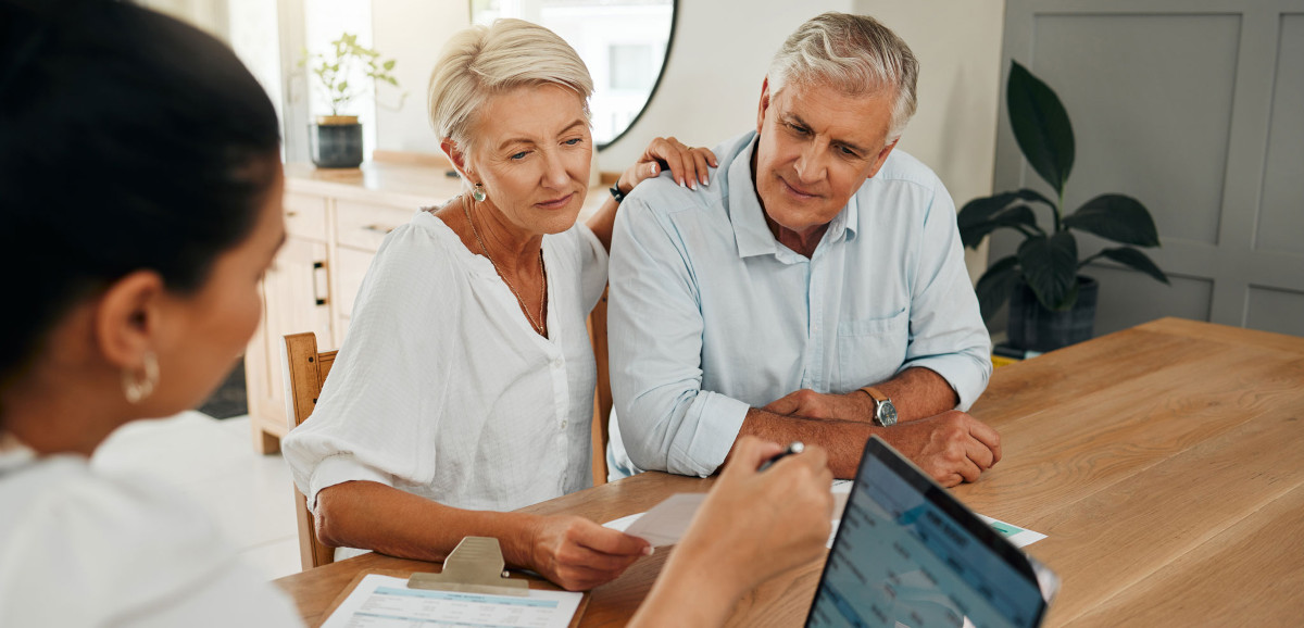 An elderly couple sitting at their kitchen table discussing a contract with a financial adviser.