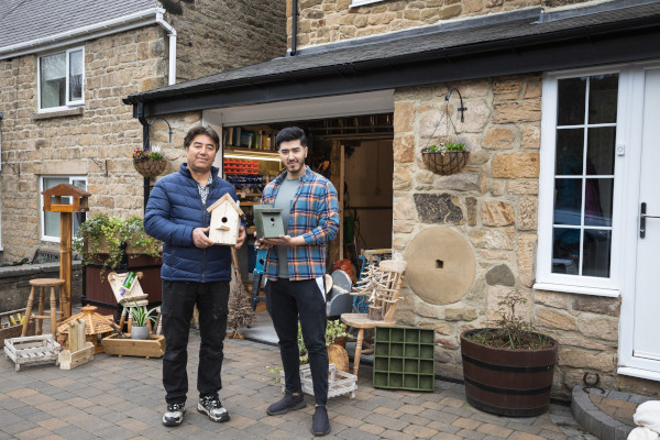 Two people standing in front of a garage of a home holding homemade bird boxes they're selling